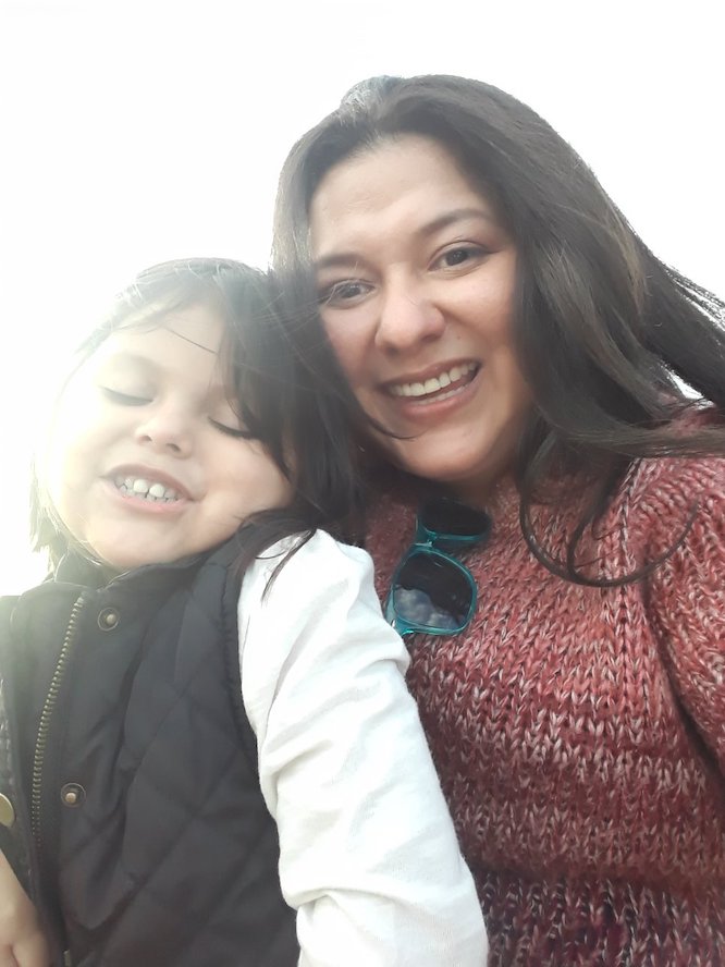 Isabel Torres smiles at the camera wearing a red sweater while her daughter sits on her lap smiling as well.