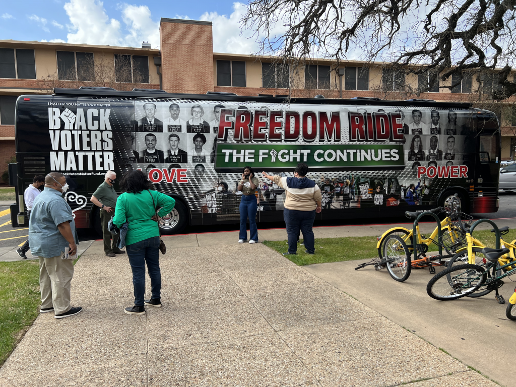 The Black Voters Matter tour bus parked at Huston-Tillotson University. There a handful of people standing outside the bus and taking photos of and with the bus in the background.