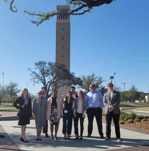 A group of students stand out side and smile for a picture in front of a tall clock tower.