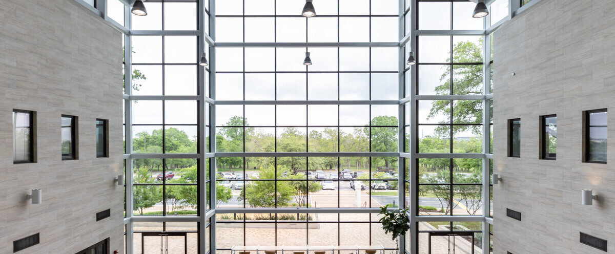 A spacious, modern lobby with a high ceiling and large floor-to-ceiling windows that provide a view of the outdoors. The interior features light-colored walls and dark flooring. Several seating areas with chairs and tables are arranged neatly. Outside, trees and a parking lot are visible through the windows. The space is well-lit with natural light streaming in.