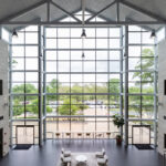 A spacious, modern lobby with a high ceiling and large floor-to-ceiling windows that provide a view of the outdoors. The interior features light-colored walls and dark flooring. Several seating areas with chairs and tables are arranged neatly. Outside, trees and a parking lot are visible through the windows. The space is well-lit with natural light streaming in.