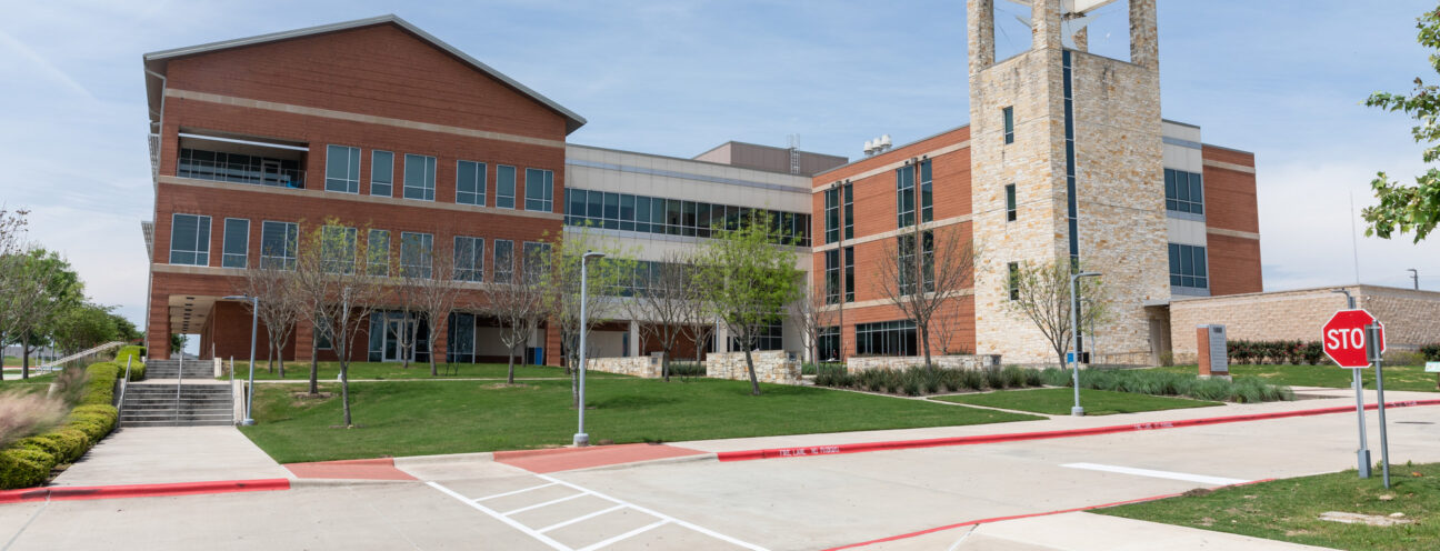 A modern educational building with a combination of brick and stone architecture. The structure features large windows and a prominent clock tower. In the foreground, there is a paved area with a stop sign and landscaped greenery, including small trees and grass. The sky is clear with a few clouds.