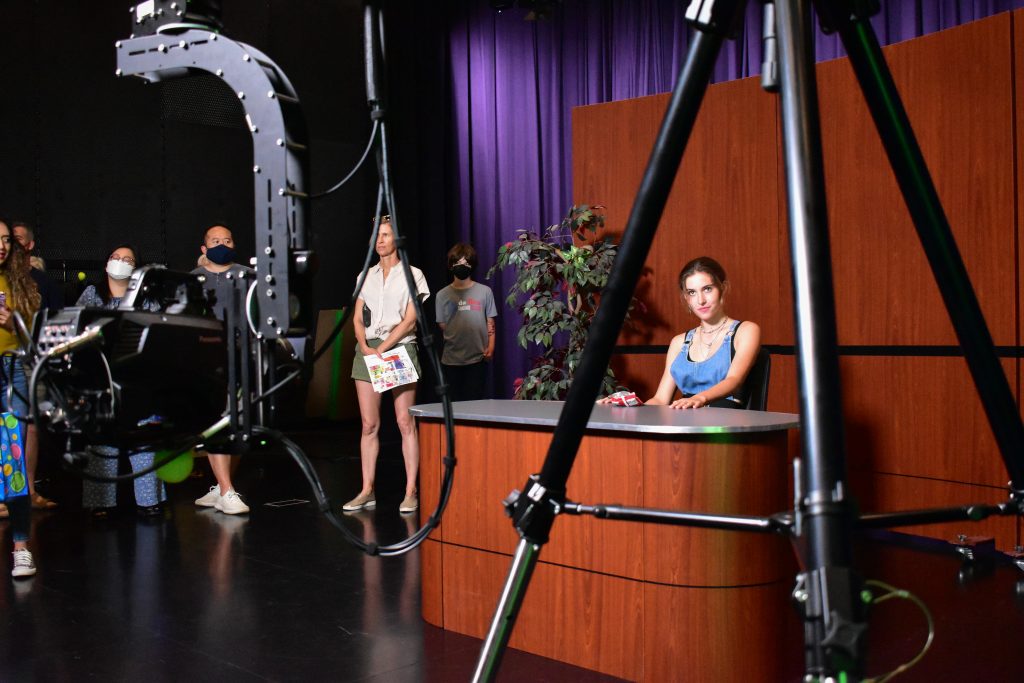 A woman sits in a news set while a camer moves in front of her.