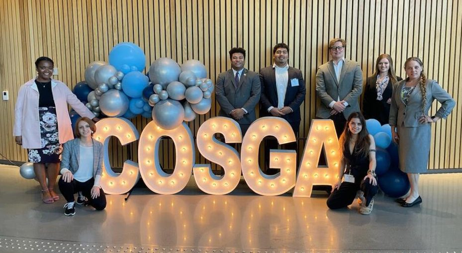 A group of students stand inside and smile for a picture next to large letters lit up in lights that spell "COSGA."