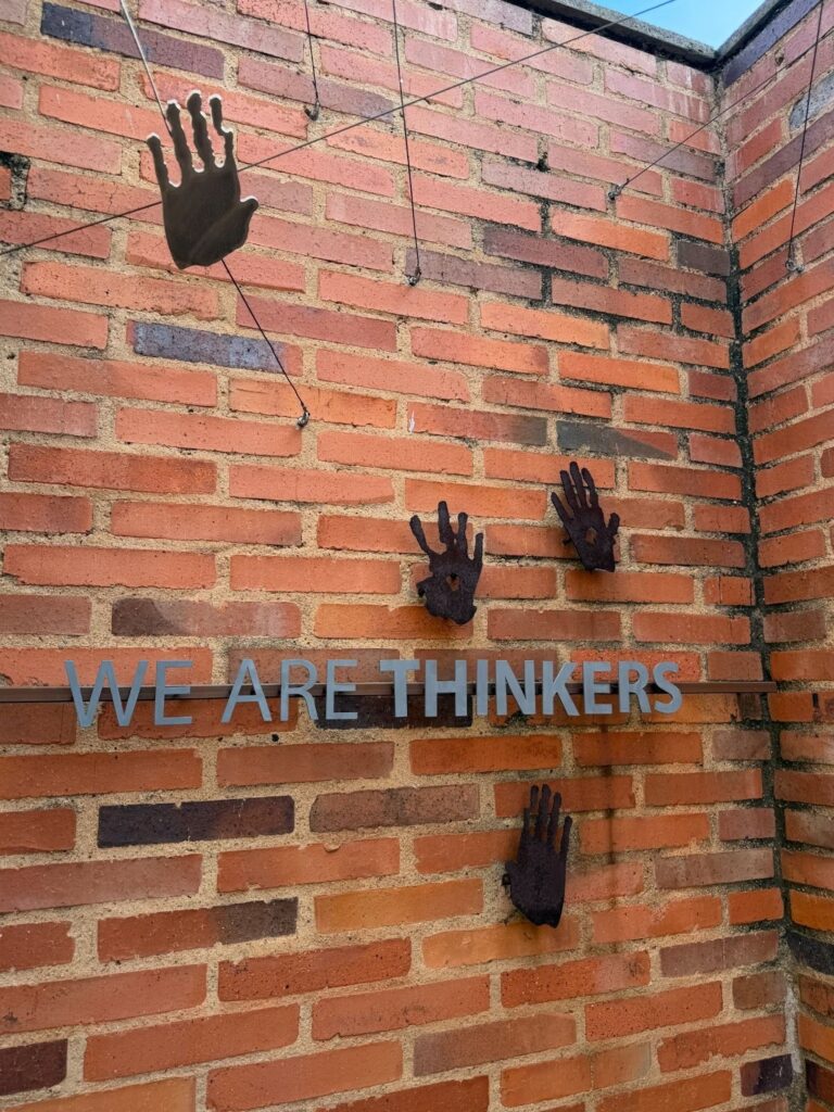 A vertical photograph of a brick wall inside the Rock Art Enclosures at the Apartheid Museum. The wall features several dark, raised handprints of varying sizes positioned at different heights. Below the handprints, the words "WE ARE THINKERS" are inscribed in blue capital letters. Natural light shines down from an opening in the ceiling above.