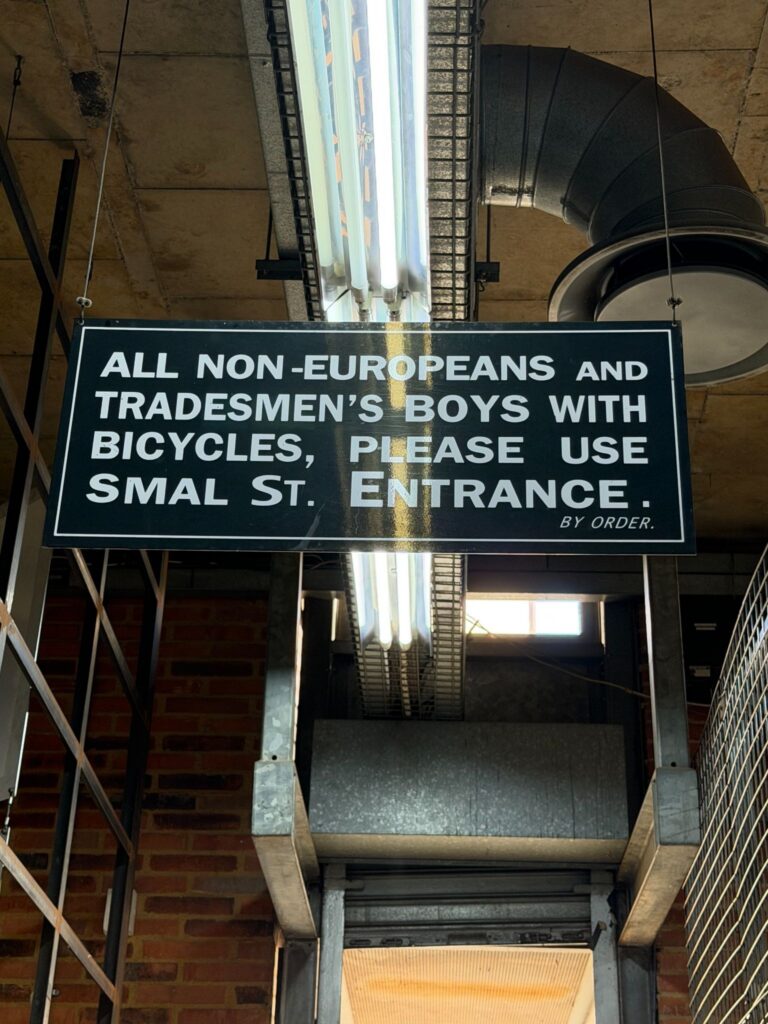A blue historical sign from South Africa's apartheid era that reads: "ALL NON-EUROPEANS AND TRADESMEN'S BOYS WITH BICYCLES, PLEASE USE SMAL ST. ENTRANCE. BY ORDER