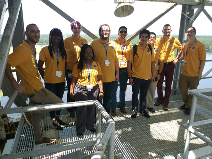 A group of students in yellow NASA polo shirts stand on a metal platform outdoors at an industrial or launch facility, smiling with safety railings and equipment visible around them.