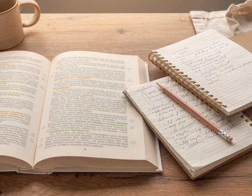 Student writing in a notebook with an open textbook nearby