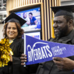 Two graduates pose with pom poms and a riverbat flag