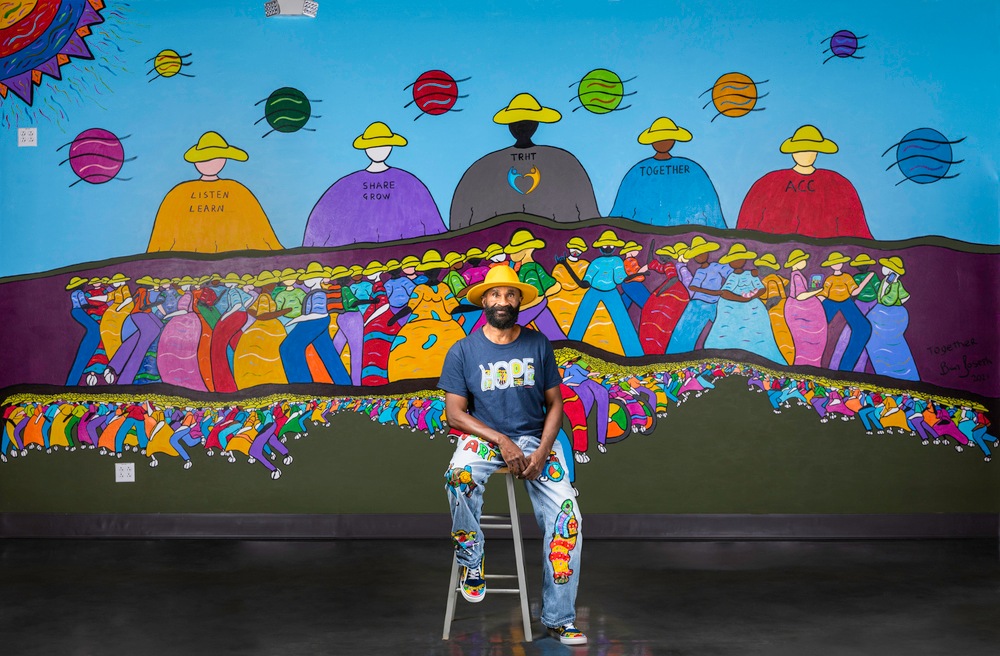 Artist Brian Joseph seated on stool in front of colorful mural he painted for ACC Truth, Racial Healing & Transformation Campus Center