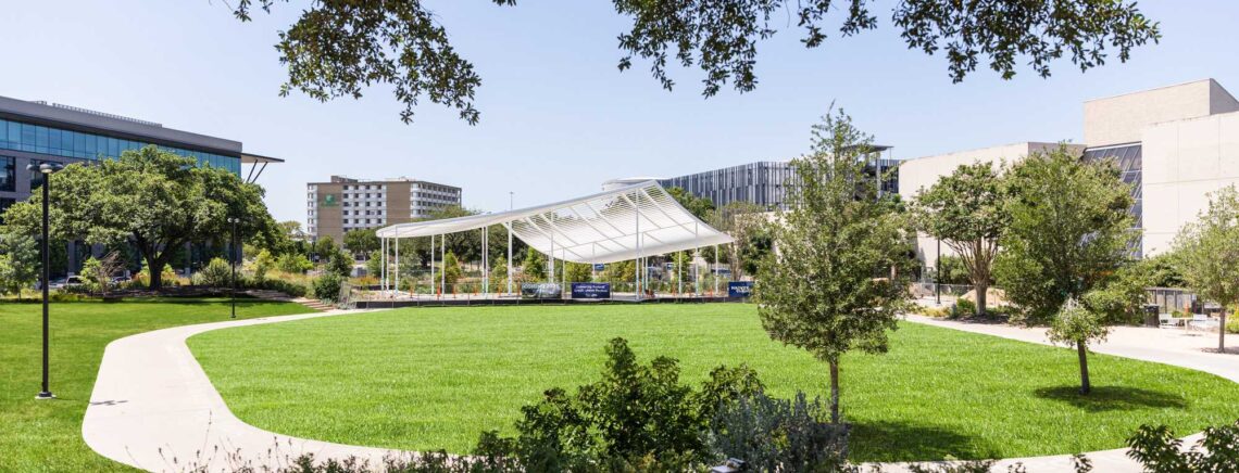 A vibrant green field with trees scattered throughout, showcasing the UFCU pavillion in the distance against a bright backdrop.