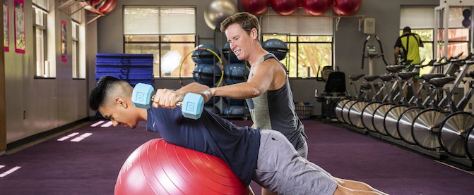 Fitness trainer assists male with large exercise ball and hand weight