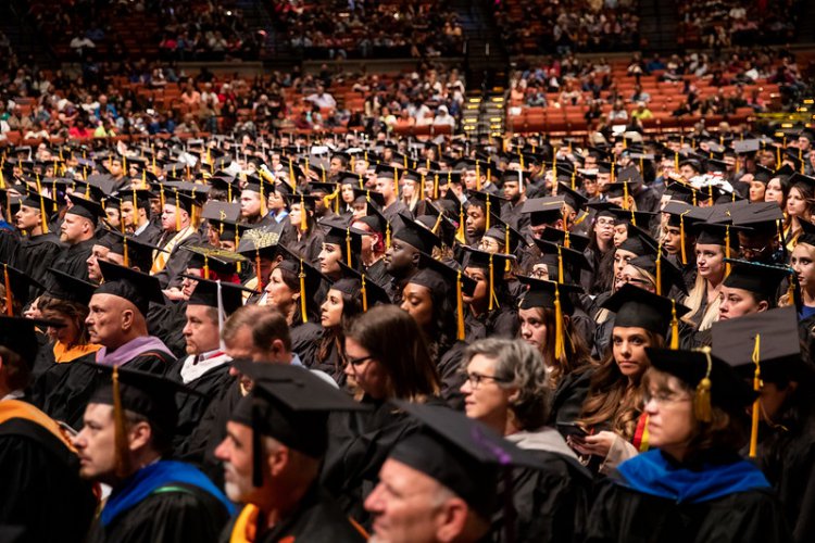 Dozens of graduates wearing caps and gowns at commencement ceremony with spectators in balcony seats in the background