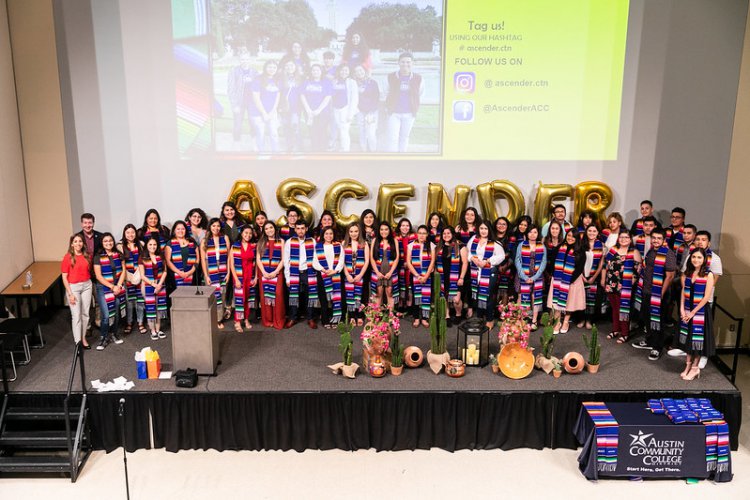 Graduates with colorful stoles in front of projected slide and gold balloons spelling Ascender