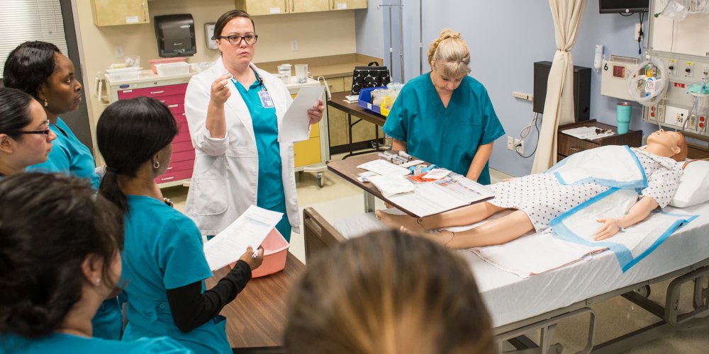 Nursing instructor talks with students in simulation lab at bedside of simulation mannequin on hospital bed