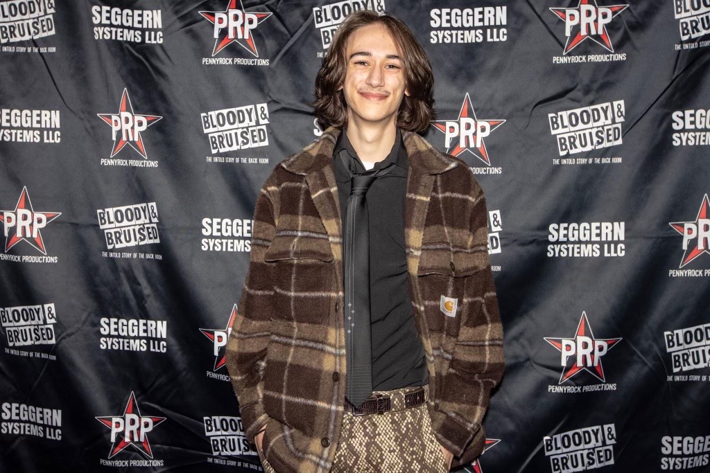 male student with long brown hair and brown plaid jacket with black shirt stands in front of a backdrop and smiles at the camera