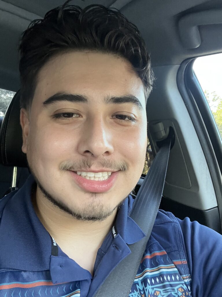 Photo of Jose, Hispanic Male with brown hair wearing a blue shirt is sitting in his car and smiling at the camera