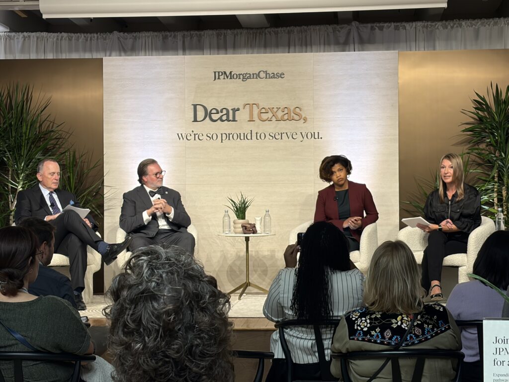 ACC Chancellor Dr. Russell Lowery-Hart sits with a panel. To his left is an older white man in a suit. On his other side is a black woman  in red shirt. Next to her is a woman in all black reading from a cards and holding a microphone.