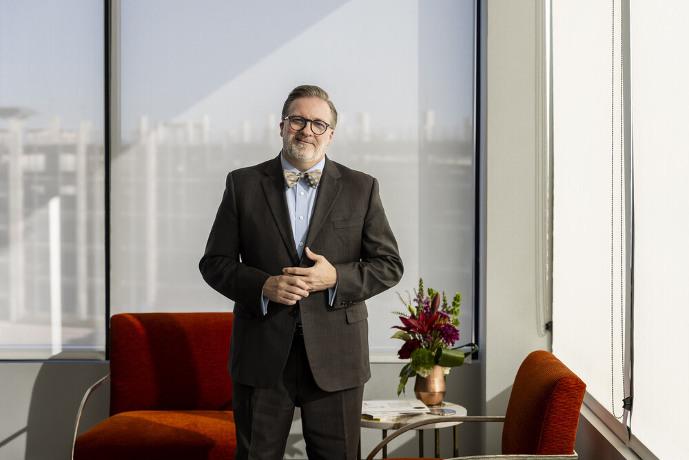 ACC Chancellor Dr. Russell Lowery-Hart, a middle-aged white man stands confidently in a modern office setting. He is wearing a dark suit with a bow tie and glasses, smiling warmly. Behind him, large windows let in natural light, revealing a cityscape outside. In the foreground, there are two orange chairs and a small table with a vase of flowers, adding a touch of color to the professional environment.