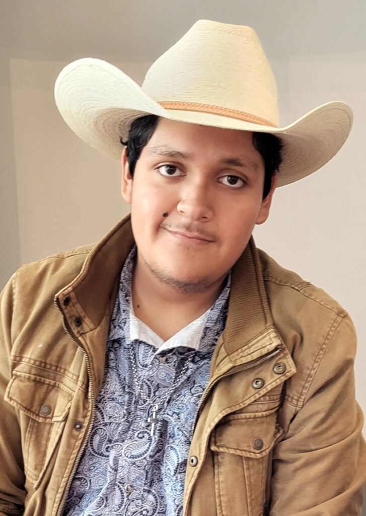 Hispanic student wearing a blue shirt and tan jacket smiles at the camera. He has short black hair and is wearing a cowboy hat in front of a neutral background