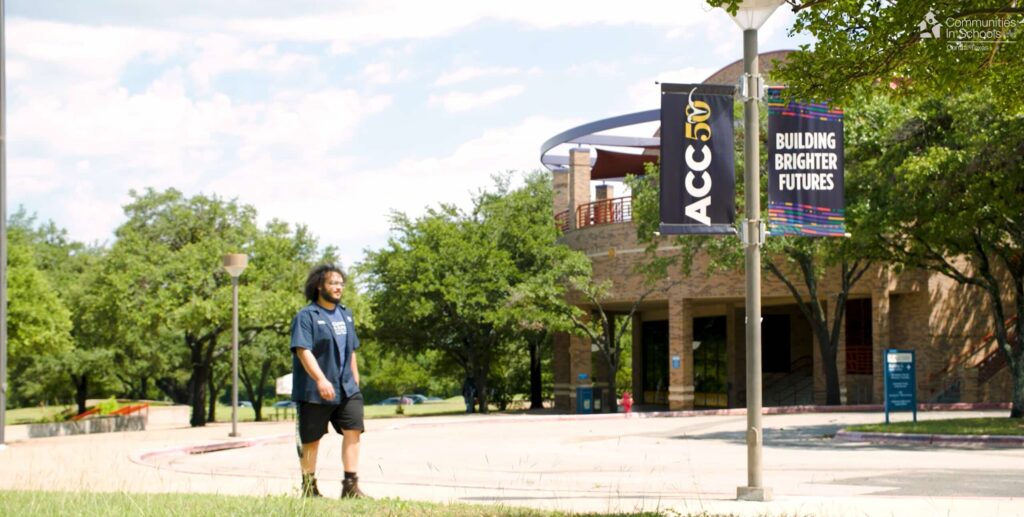 Male student walking outside the Riverside Campus. His is wearing dark shorts and an ACC button up. He has curly hair. Ahead of him is a light pole with ACC banners. Behind him is a campus and trees.