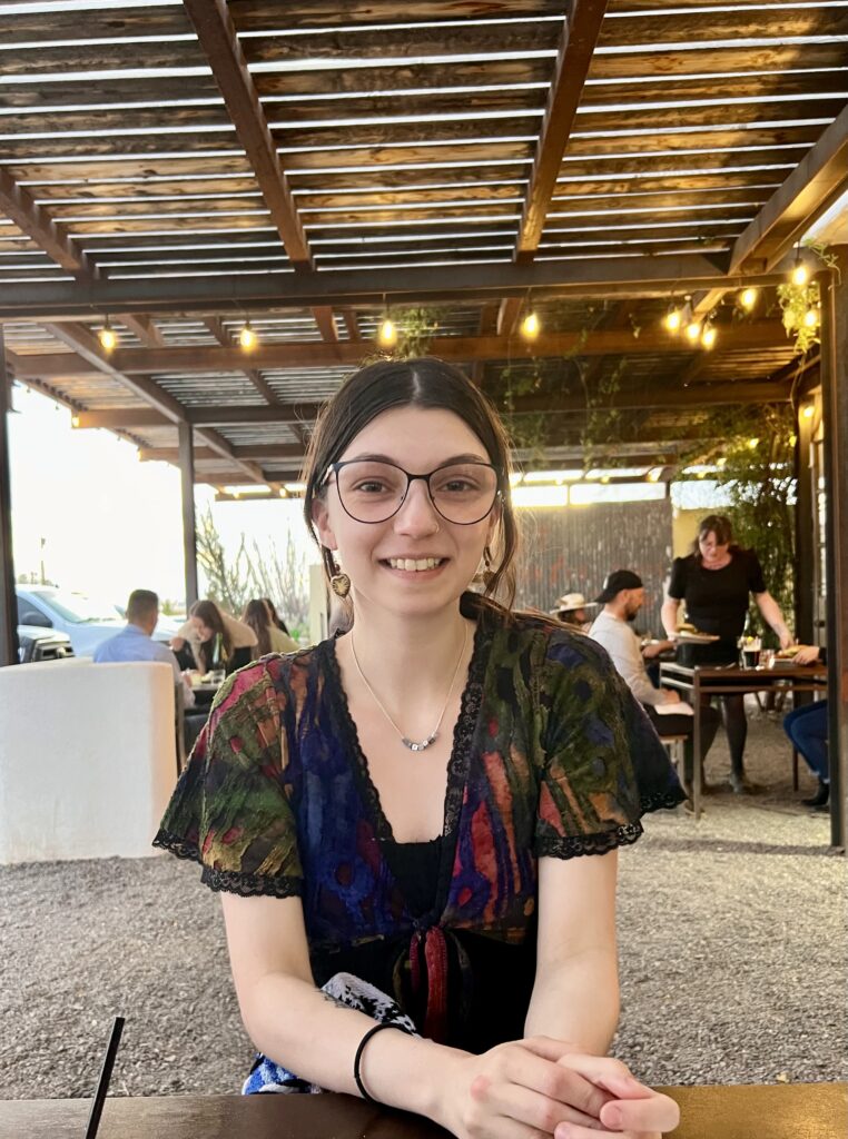 Student with multicolored dress smiles at the camera. She has brown hair that is pulled back and wearing glasses. The is sitting outside under a patio. 