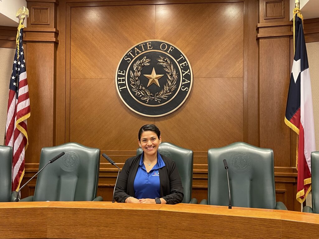 Student in blue collared shirt and black jacket sits in a green chair at the dias with two open chairs next to her. She sits in front of the seal of Texas with an American and USA flag behind her