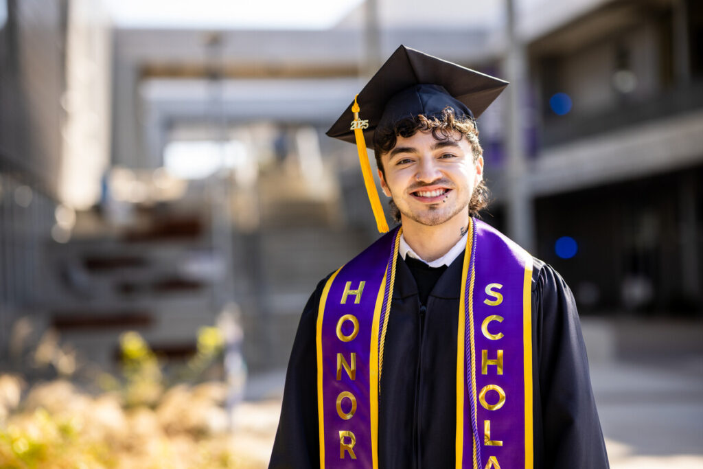 A young man stands outdoors, smiling proudly while wearing a black graduation gown and cap. He has curly hair and is adorned with a purple and gold honor cord and sash that reads "HONOR SCHOLAR." The background features a modern architectural setting with blurred structures and greenery.