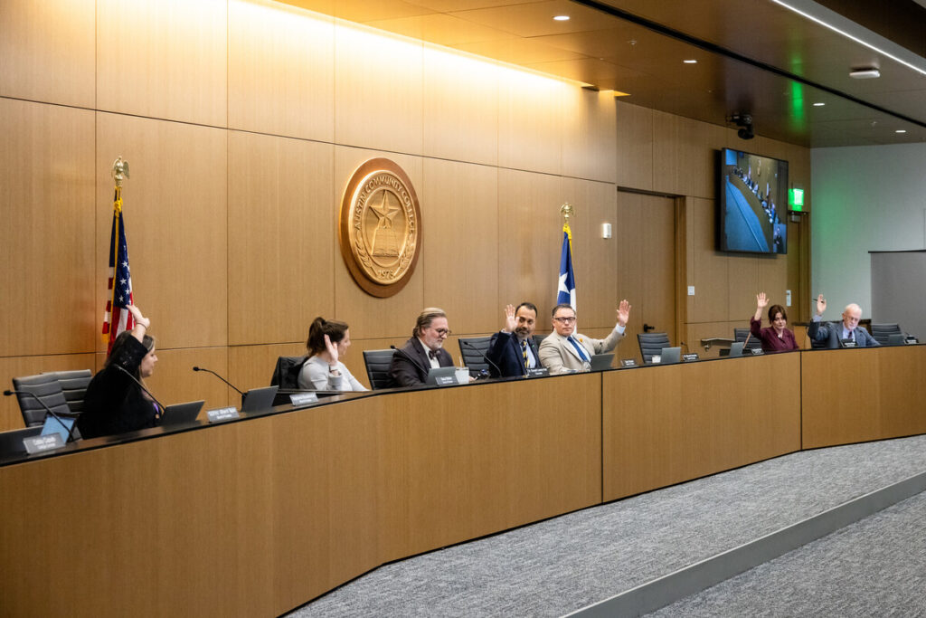 An ACC District Board of Trustees regular meeting is taking place in a modern conference room. Six trustees and the chancellor are seated at a dais, with some raising their hands in a voting gesture. The room features a large wooden wall with an ACC District seal, and there are American and Texas flags displayed. A screen in the background shows a video feed of the meeting. The atmosphere appears formal and organized.