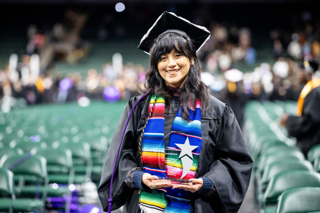 A smiling graduate stands in a large auditorium, wearing a black graduation gown and cap. She holds a trophy in her hands and is draped with a colorful, striped graduation stole. The background features blurred figures of other graduates and empty seats, indicating a graduation ceremony.