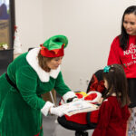 A woman dressed as an elf in a green costume hands a gift to a young girl in a red dress. The girl looks excited as she receives the gift. In the background, a woman wearing a red sweater that says "On the Naughty List" watches with a smile. The setting is festive, with holiday decorations visible.