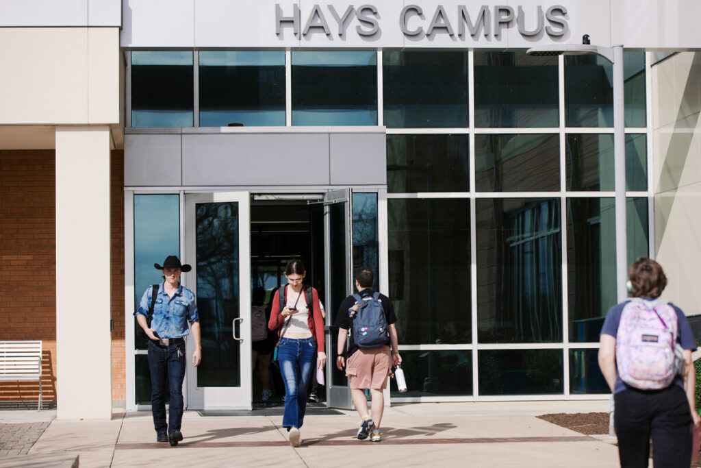 A group of four people walking out of a modern building labeled "Hays Campus." Two individuals are exiting through the main door, one wearing a cowboy hat and the other carrying a camera. A third person is walking towards the entrance, and a fourth person is seen in the background, wearing a backpack. 