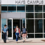 A group of four people walking out of a modern building labeled "Hays Campus." Two individuals are exiting through the main door, one wearing a cowboy hat and the other carrying a camera. A third person is walking towards the entrance, and a fourth person is seen in the background, wearing a backpack. The setting appears to be a sunny day with clear skies.