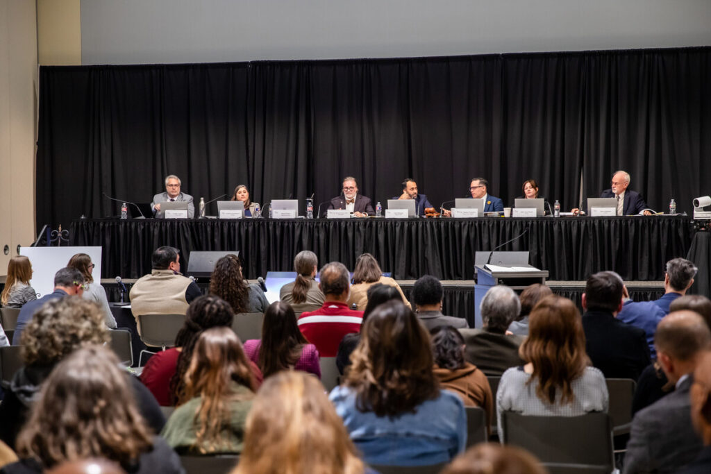 A panel of speakers is seated at a long table in front of a large audience. The panelists, consisting of seven individuals, are engaged in discussion, with microphones in front of them. The audience, composed of various individuals, is seated in rows, facing the panel. The setting appears to be a formal event, likely a conference or meeting, with a backdrop of black curtains.