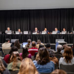 A panel of speakers is seated at a long table in front of a large audience. The panelists, consisting of six individuals, are engaged in discussion, with microphones in front of them. The audience, composed of various individuals, is seated in rows, facing the panel. The setting appears to be a formal event, likely a conference or meeting, with a backdrop of black curtains.