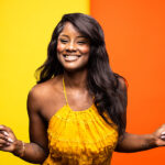 A smiling woman with long, wavy black hair is wearing a bright yellow dress with a textured, ruffled design. She stands against a vibrant background that is split between yellow and orange. Her joyful expression and playful pose convey a sense of happiness and energy.