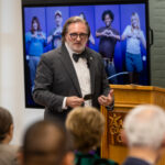 A man in a gray suit and bow tie stands at a wooden podium, speaking to an audience. Behind him is a large screen displaying images of diverse individuals making heart shapes with their hands. The setting appears to be a conference or community event, with attendees seated and facing the speaker.