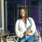 A young woman with long, braided hair smiles brightly while holding a laptop and a notebook. She is wearing a white sweater and standing in a modern, well-lit space with large windows. In the background, another person is seated at a table, focused on writing or studying. The environment features contemporary furniture and a vibrant atmosphere.