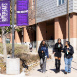 Three young women walk together along a pathway outside a building. They are dressed in casual attire, with one wearing a black dress and knee-high boots. The group appears to be engaged in conversation, each holding a drink. In the background, there are other people walking and a banner that reads "RIVERBATS YOU BELONG START HERE." The scene is set in a campus-like environment with trees and a brick building.