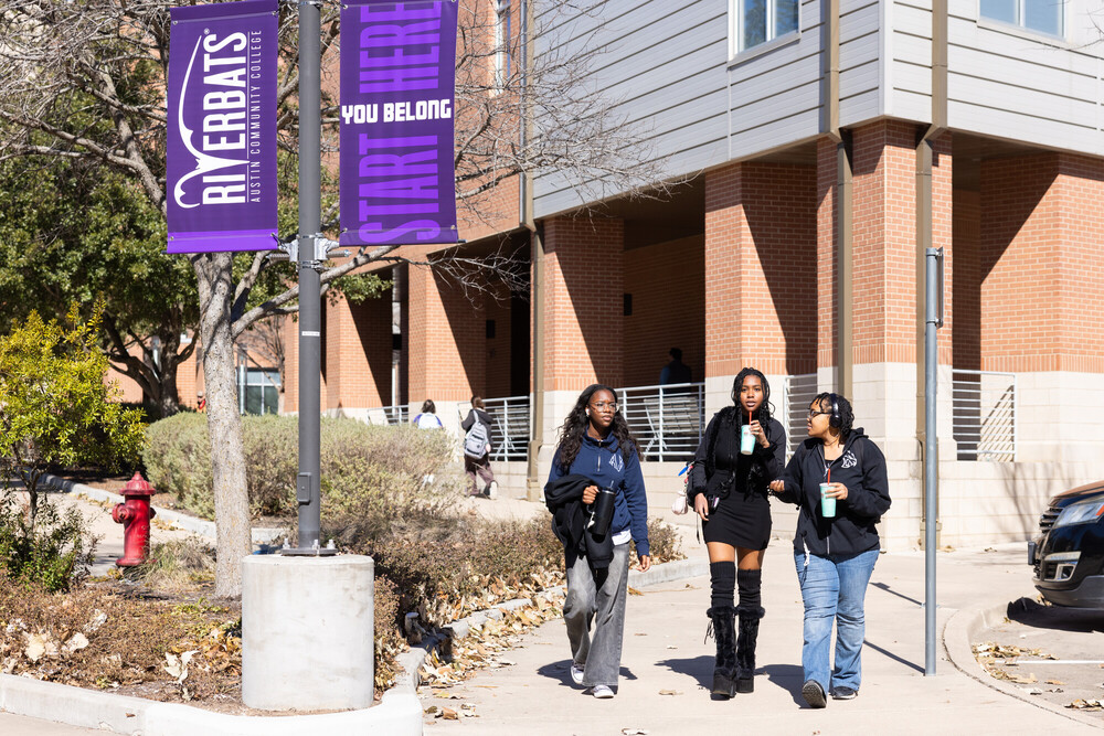 Three young women walk together along a pathway outside a building. They are dressed in casual attire, with one wearing a black dress and knee-high boots. The group appears to be engaged in conversation, each holding a drink. In the background, there are other people walking and a banner that reads "RIVERBATS YOU BELONG START HERE." The scene is set in a campus-like environment with trees and a brick building.