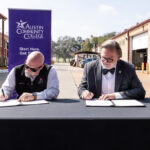 Two men are seated at a table, signing documents. They are in an outdoor setting with a banner in the background that reads "Austin Community College District" and the phrase "Start Here. Get You." One man is wearing a black vest and glasses, while the other is dressed in a suit and bow tie. The school environment is a facility with industrial elements visible in the background.