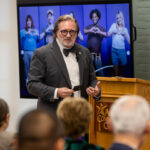 A man in a gray suit and bow tie stands at a wooden podium, speaking to an audience. Behind him is a large screen displaying images of diverse individuals making heart shapes with their hands. The setting appears to be a conference or community event, with attendees seated and facing the speaker.