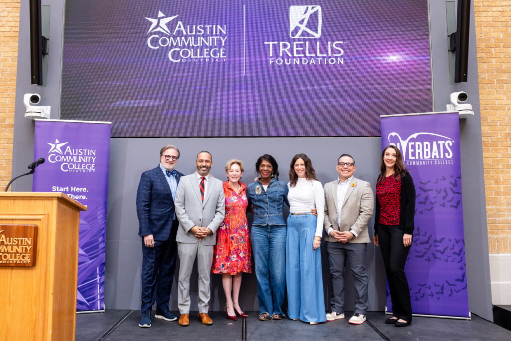 A group of seven individuals stands on a stage in front of a large screen displaying the logos of Austin Community College and the Trellis Foundation. The setting appears to be a formal event, with a podium on the left. The individuals are dressed in a mix of professional and casual attire, smiling and posing together. Banners on either side of the stage. The atmosphere is celebratory and engaging.