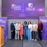 A group of seven individuals stands on a stage in front of a large screen displaying the logos of Austin Community College and the Trellis Foundation. The setting appears to be a formal event, with a podium on the left. The individuals are dressed in a mix of professional and casual attire, smiling and posing together. Banners on either side of the stage. The atmosphere is celebratory and engaging.