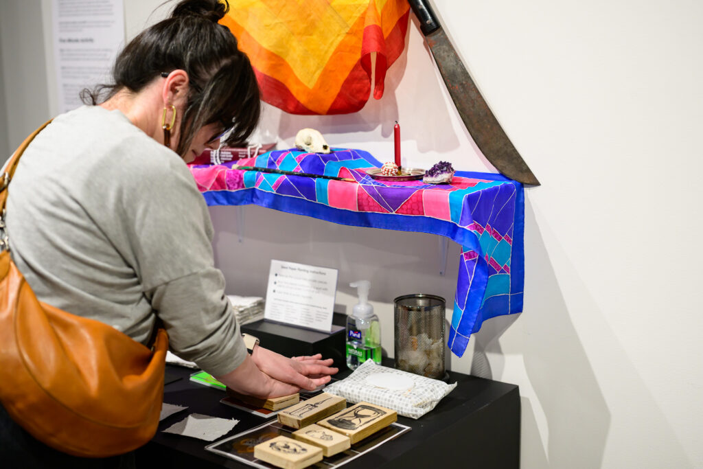 A woman in a grey shirt with a tan purse is looking at an art display. She is interacting by making a stamp. 