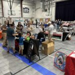 multiple rows of tables with information on them. People walk around looking at them. They look to be inside a school gym