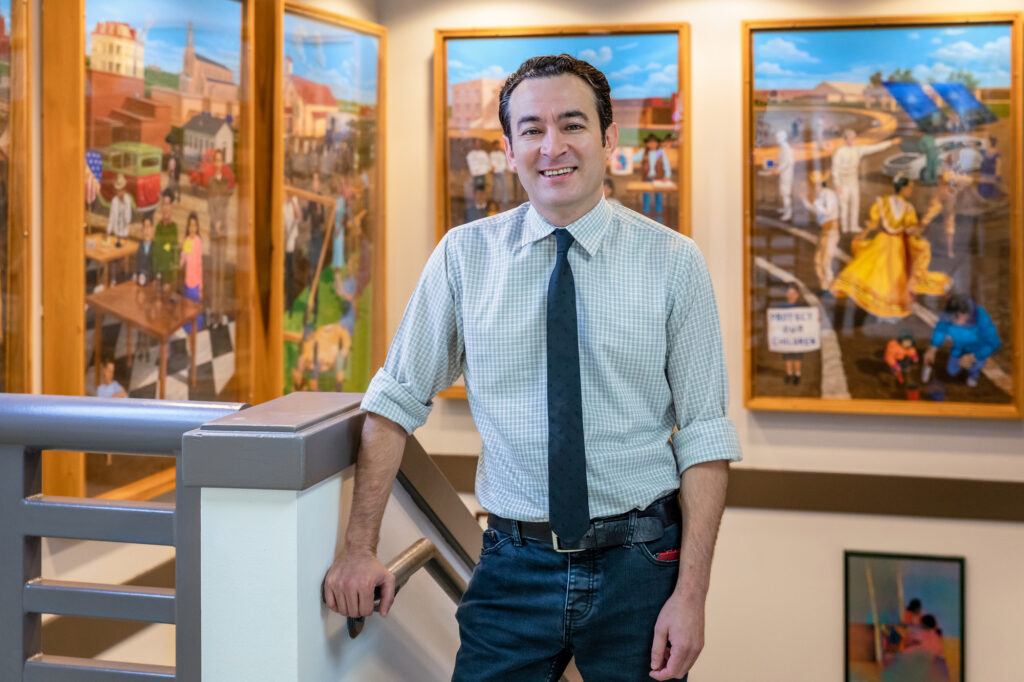 A man stands casually at the top of a staircase, leaning against a railing. He has dark hair and is wearing a light checkered shirt with a tie. Behind him, colorful paintings depicting various scenes are displayed on the walls, showcasing vibrant artwork that reflects cultural themes. The setting appears to be an indoor space with good lighting.