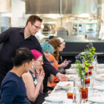 A server in a black shirt is presenting a dish to a group of diverse diners seated at a long table. The table is elegantly set with white plates, glasses of iced tea, and small vases of flowers. The diners, including individuals with pink and blue hair, are engaged in conversation and using their phones, creating a lively atmosphere. In the background, a kitchen can be seen, indicating a restaurant setting.