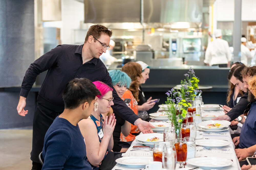 A server in a black shirt is presenting a dish to a group of diverse diners seated at a long table. The table is elegantly set with white plates, glasses of iced tea, and small vases of flowers. The diners, including individuals with pink and blue hair, are engaged in conversation and using their phones, creating a lively atmosphere. In the background, a kitchen can be seen, indicating a restaurant setting.