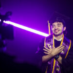 A smiling graduate stands proudly in a black cap and gown, adorned with purple and gold honor cords. The background features vibrant purple lighting, creating a celebratory atmosphere. The graduate has curly hair and a tattoo visible on their neck, exuding a sense of joy and accomplishment.